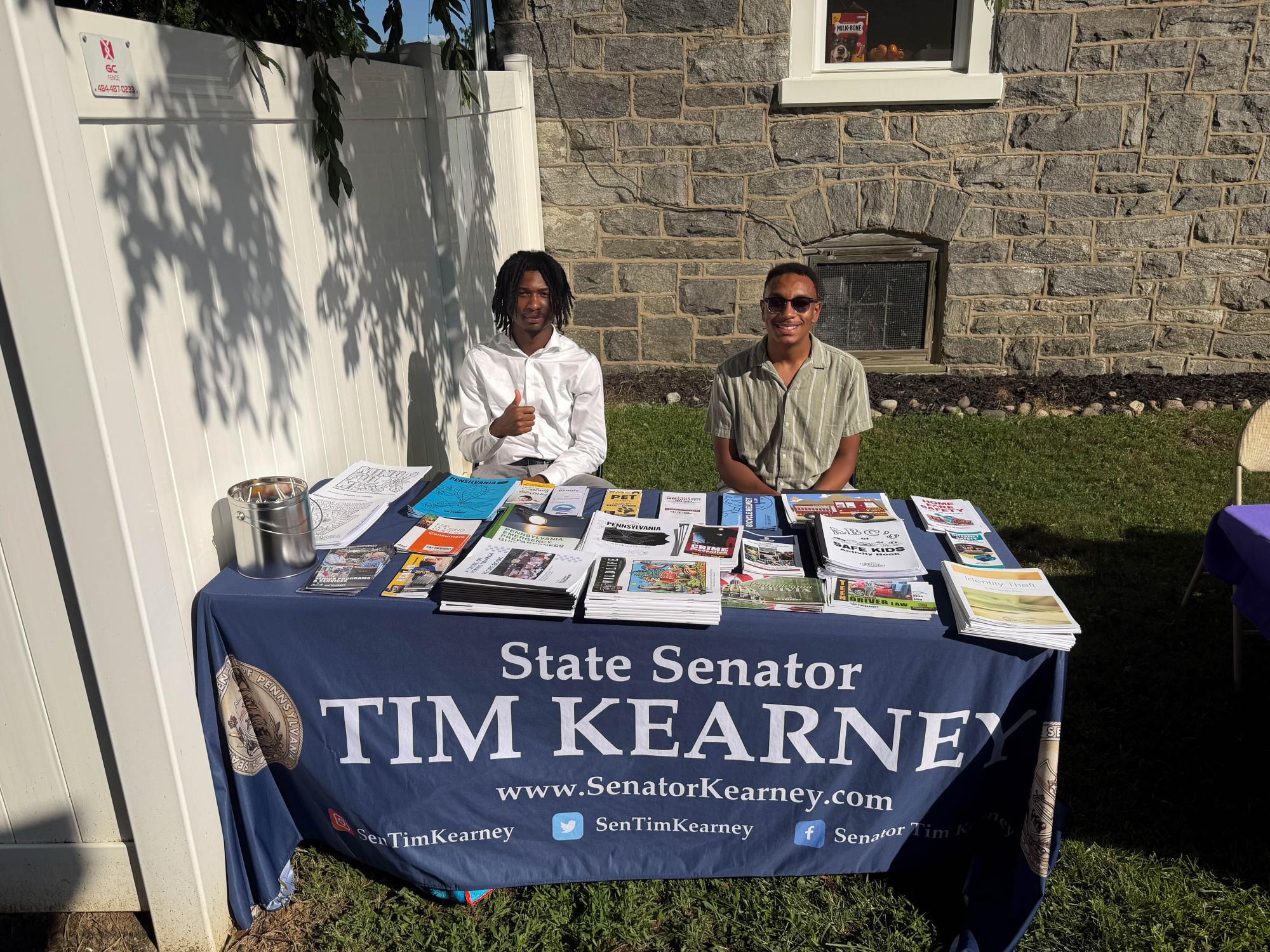 Two people sit behind an outdoor information table with pamphlets and materials. A blue tablecloth identifies the office of State Sen. Tim Kearney.