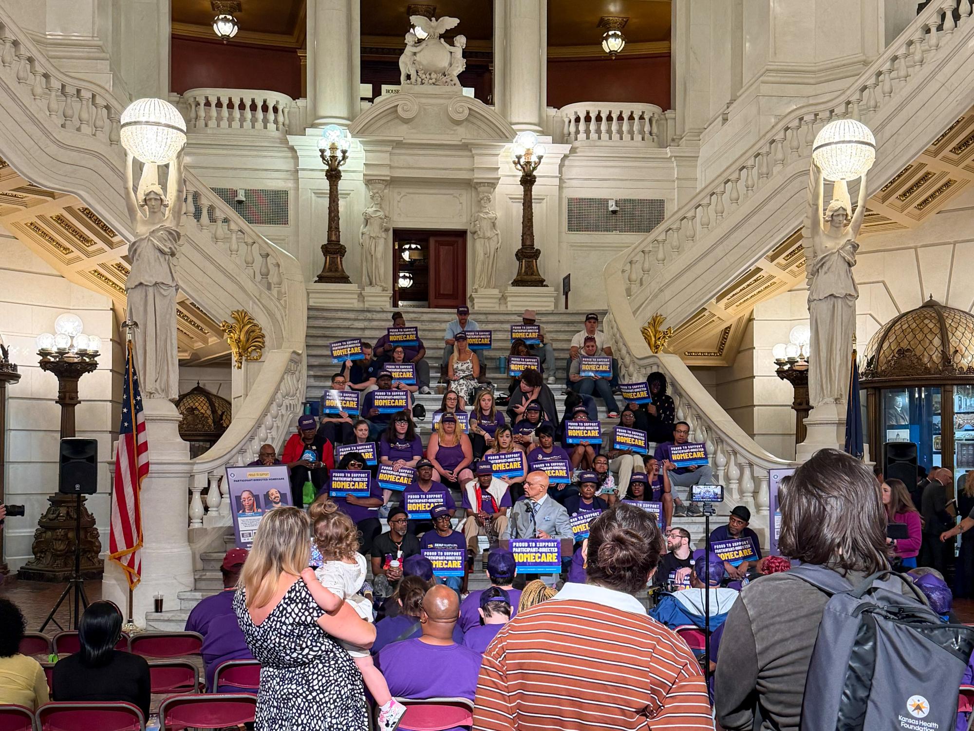 A large crowd sits and stands inside an ornate hall with marble columns, gold detailing and a grand staircase. Several people hold blue signs.