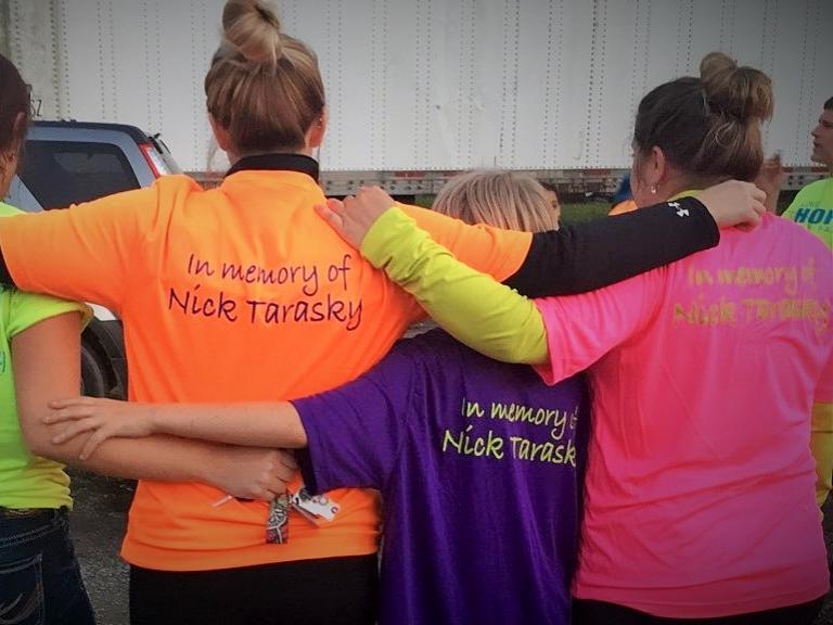 A group poses with their backs to the camera; their t-shirts read "In memory of Nick Tarasky"