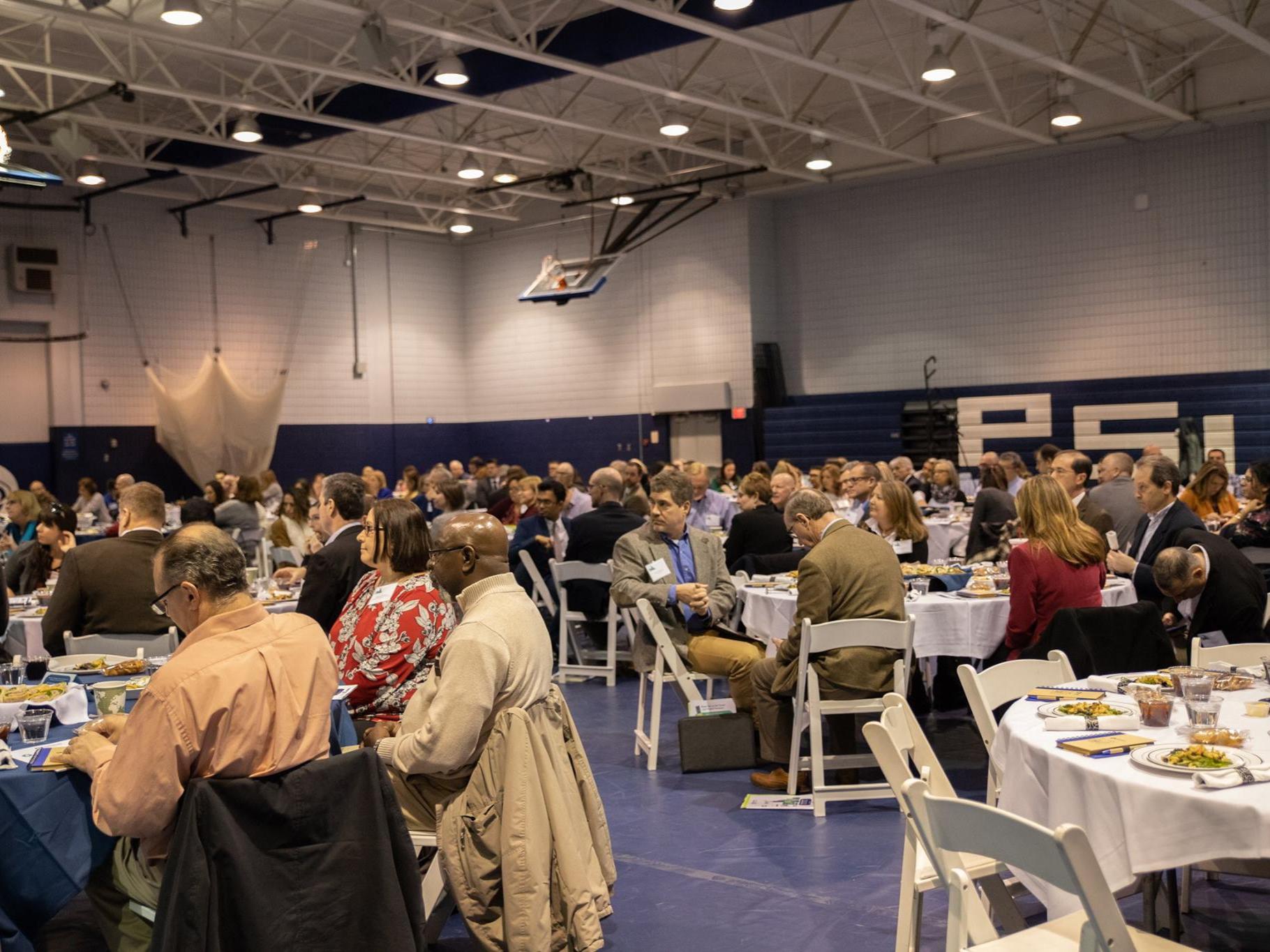 Hundreds of people sit at tables and chairs in gymnasium