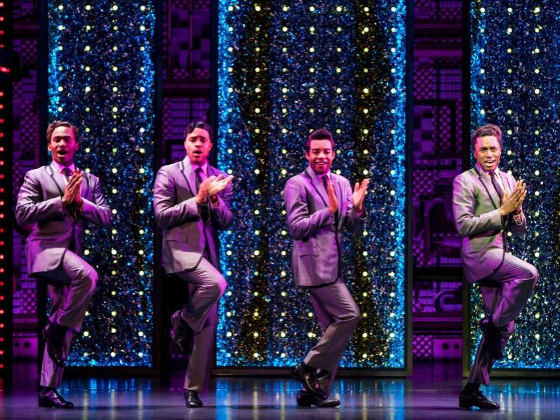 Four men wearing similar stage suits sing and dance in unison.