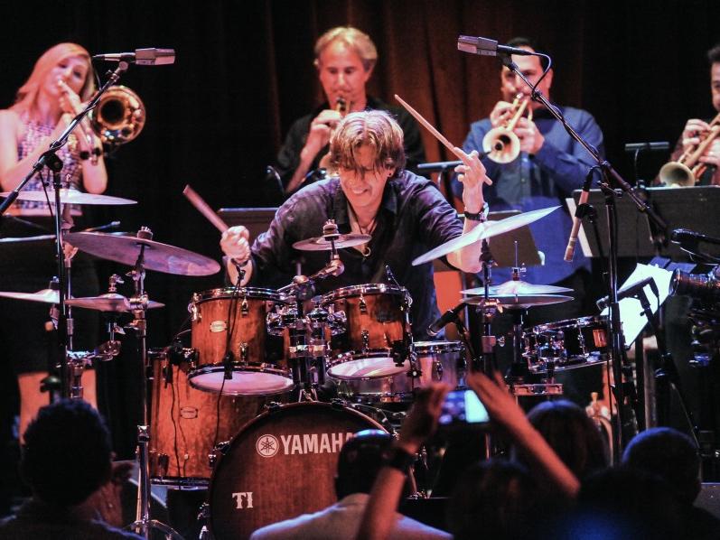 Drummer Tommy Igoe sits behind his kit and taps the cymbals while four brass musicians play behind him.