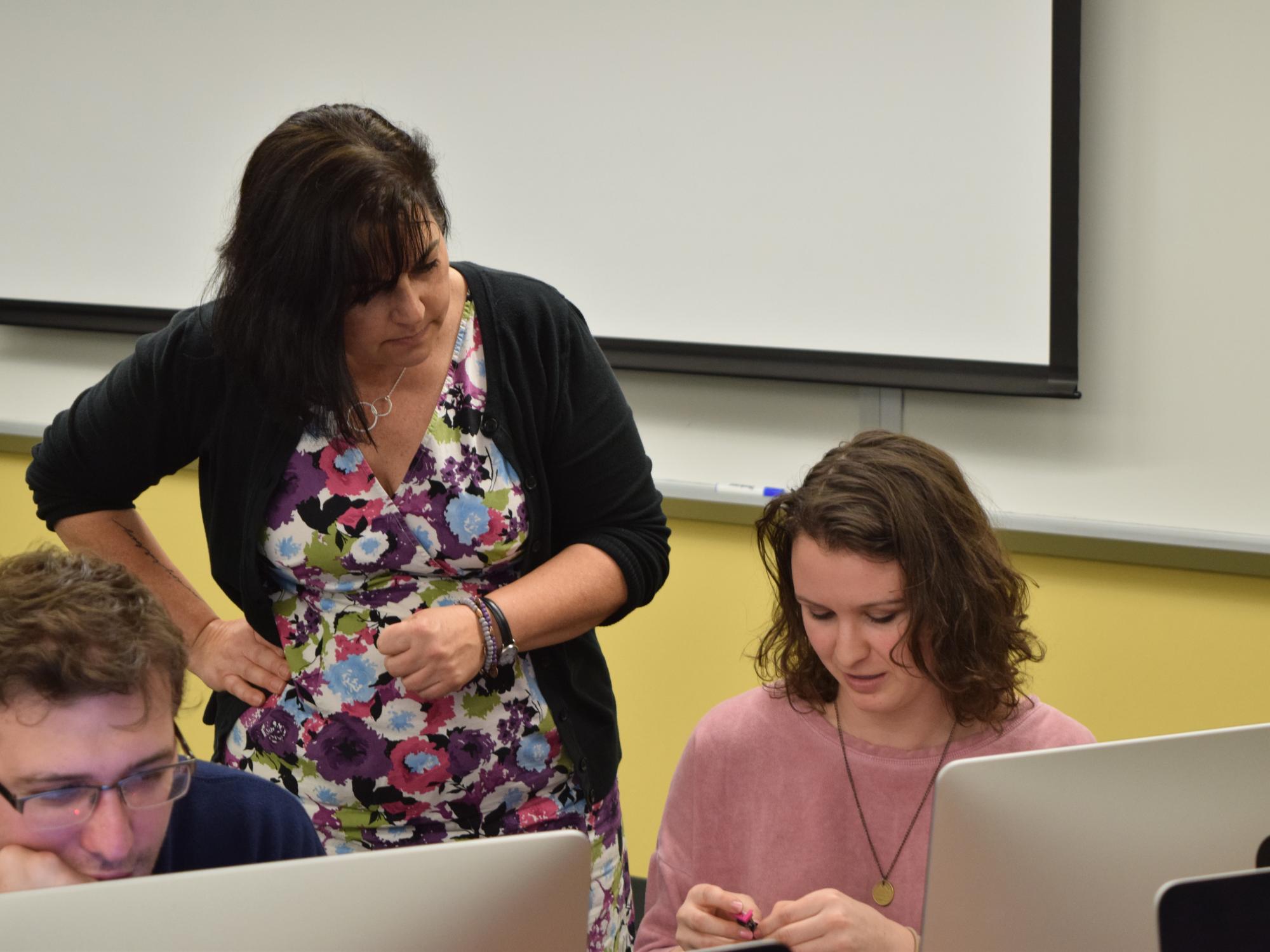 An instructor in a floral dress observes two students as they work at computers