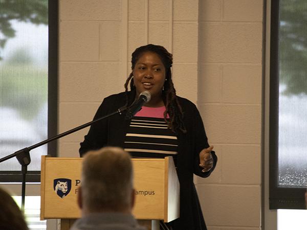 A woman in a black blazer speaks at a podium to a crowd of Penn State affiliates.