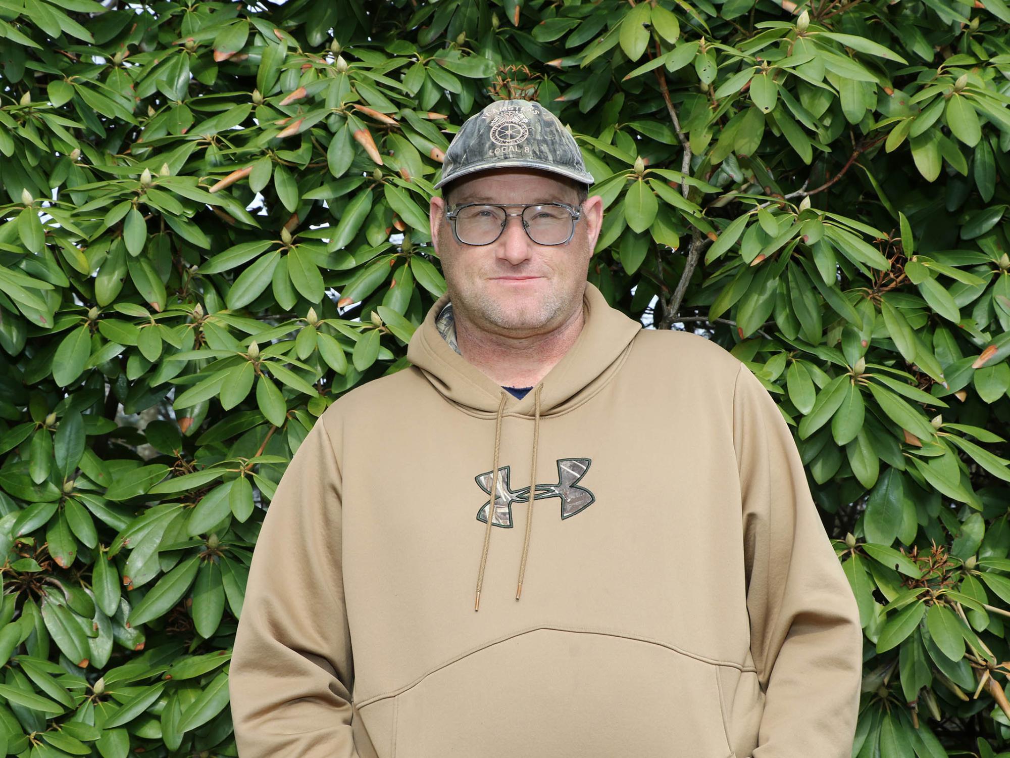 Man in hat and hooded sweatshirt standing in front of green trees.