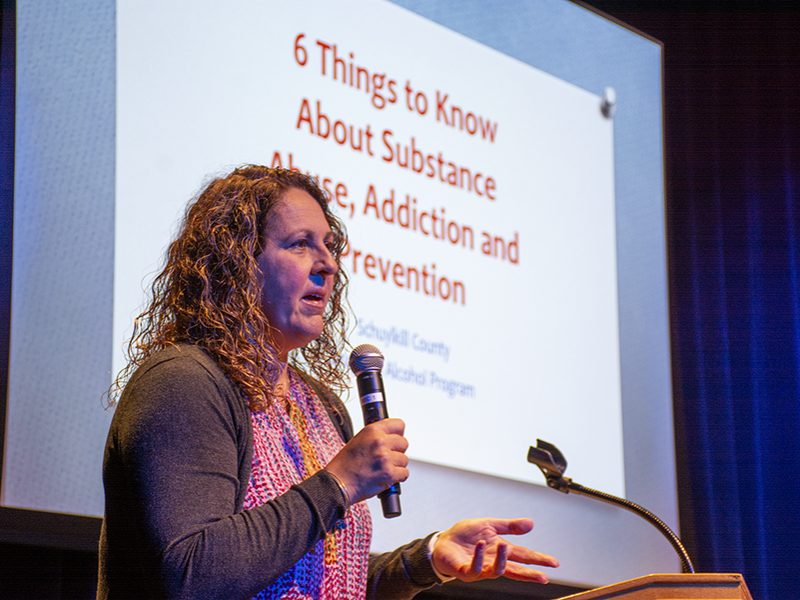 Diane Rowland holds a microphone while standing in front of a projector slide reading "Six Signs to Know about Substance Abuse, Addiction, and Prevention