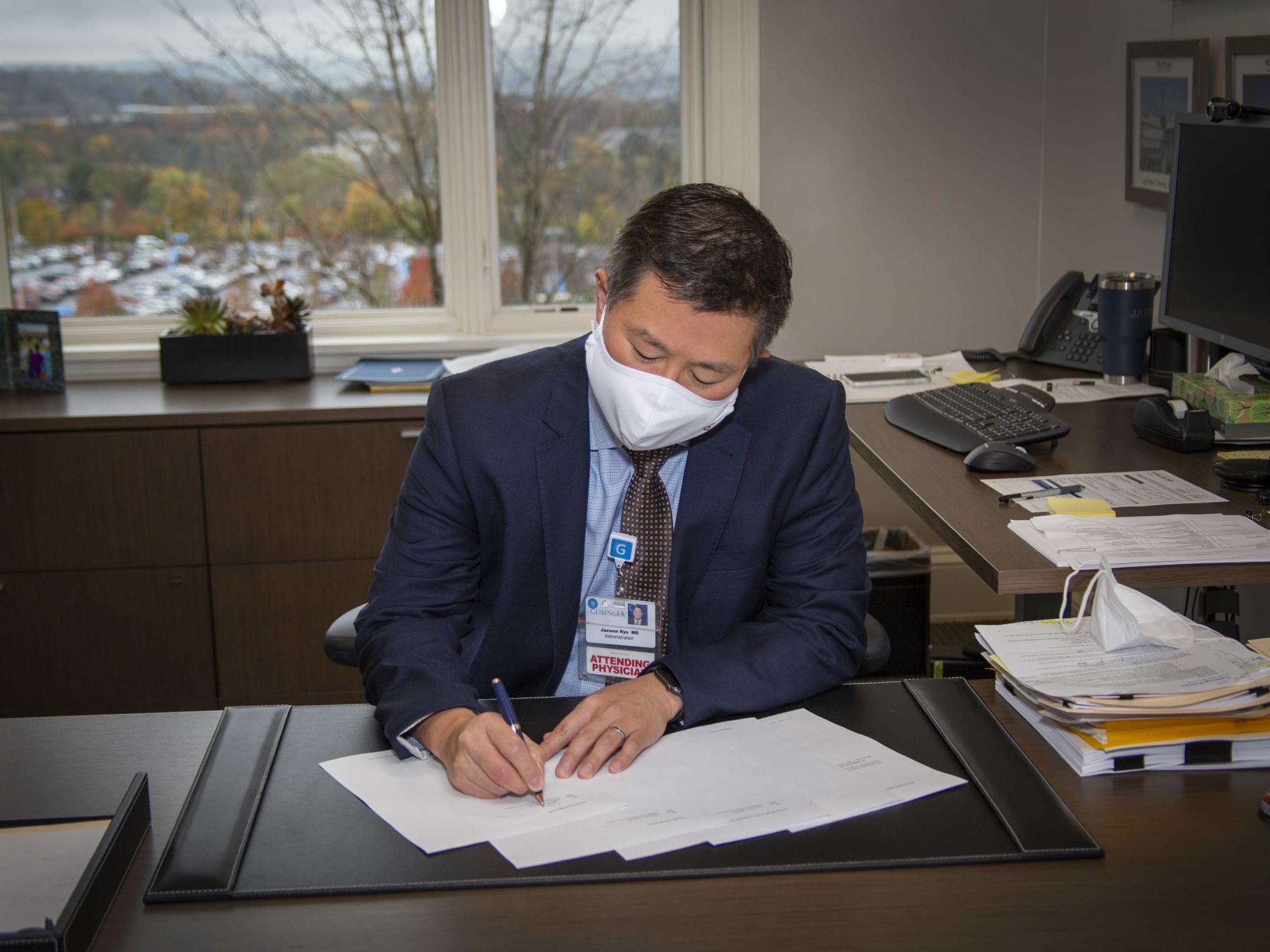 Dr. Jaewon Ryu, seated at his office desk, signs a document.