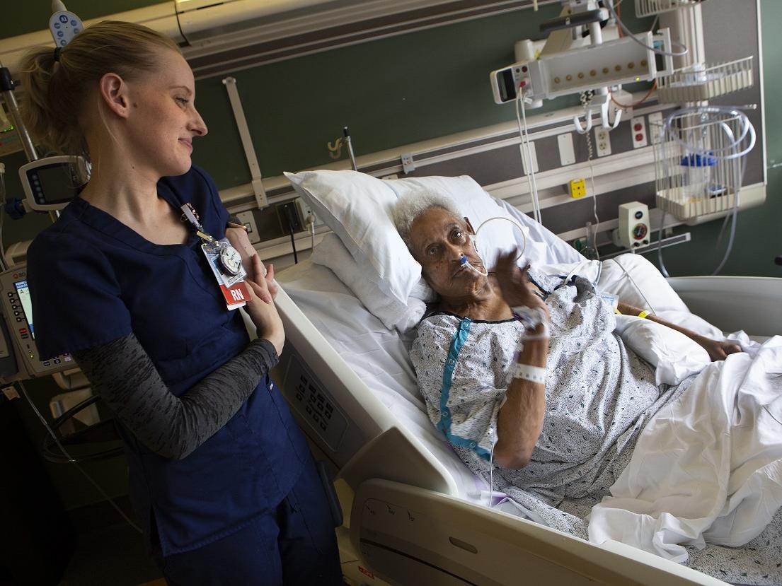 Stroke patient Gracie Banks waves from her hospital bed toward a monitor on the wall. She wears a hospital gown and is connected to tubing and equipment by her bed. Registered nurse Sara Myers smiles and leans on the hospital bed. 