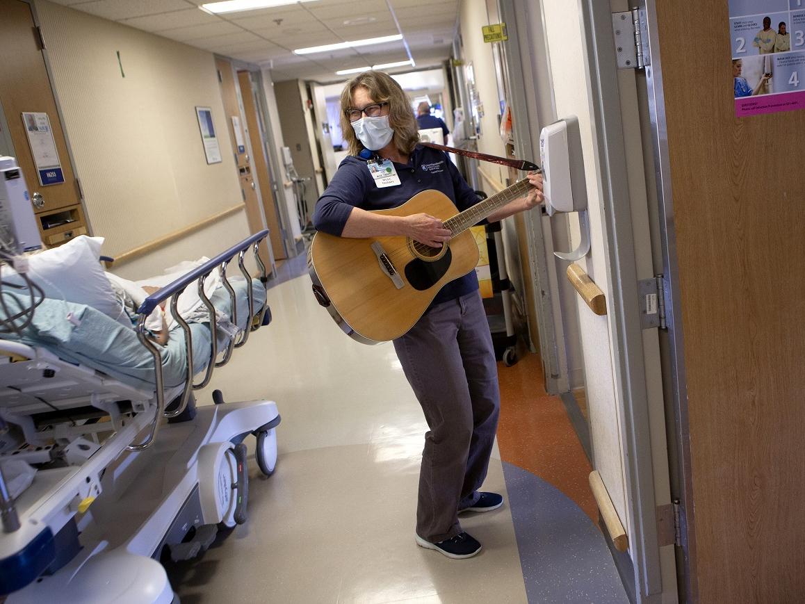 Jan Stouffer, wearing a face mask, strums a guitar and looks toward a patient on a rolling bed in the hallway of a hospital floor. She has glasses and shoulder-length wavy hair.