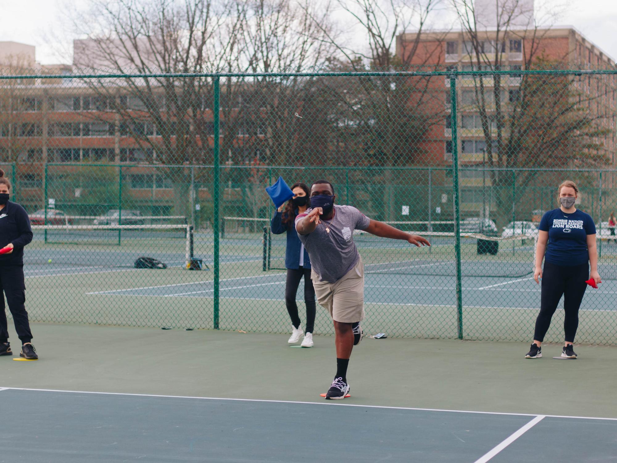 Four individuals stand on a tennis court while one tosses a bean bag.