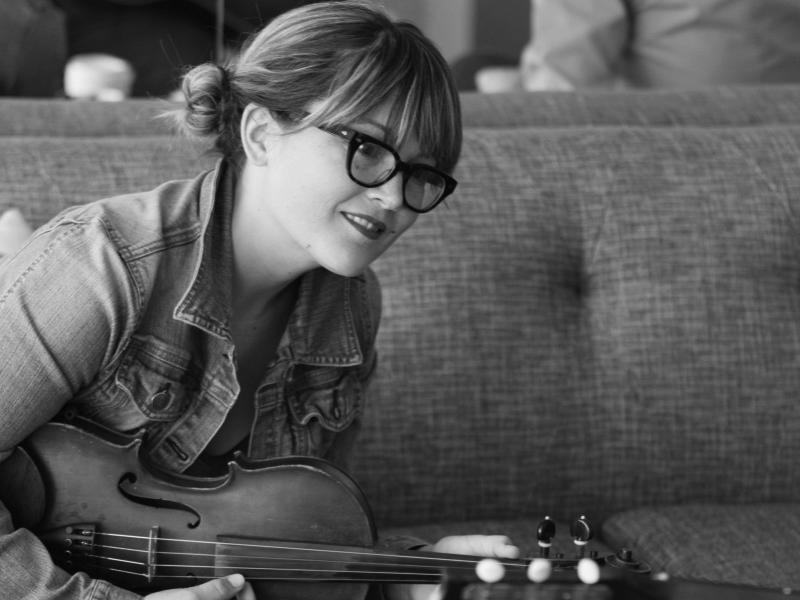 Sara Watkins sits on a couch with violin in hand and smiles.