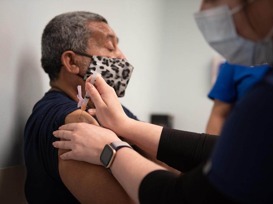 Samuel Garcia, who is seated and wears a mask, looks straight ahead as he receives a COVID-19 vaccine into his upper arm from a health care worker.