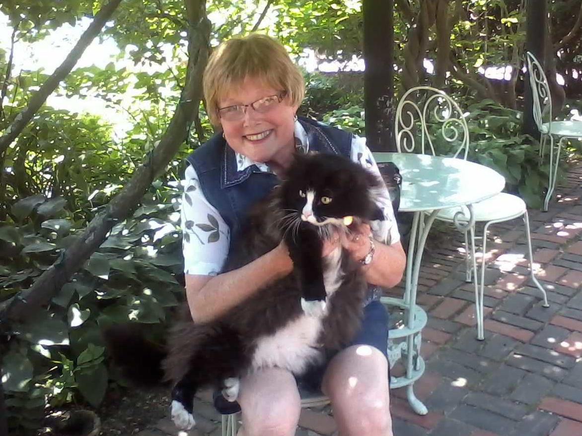 Glenna Brensinger sits on a chair on a brick patio on a sunny day, holding a cat in her lap and smiling. Trees and other vegetation are in the near background.