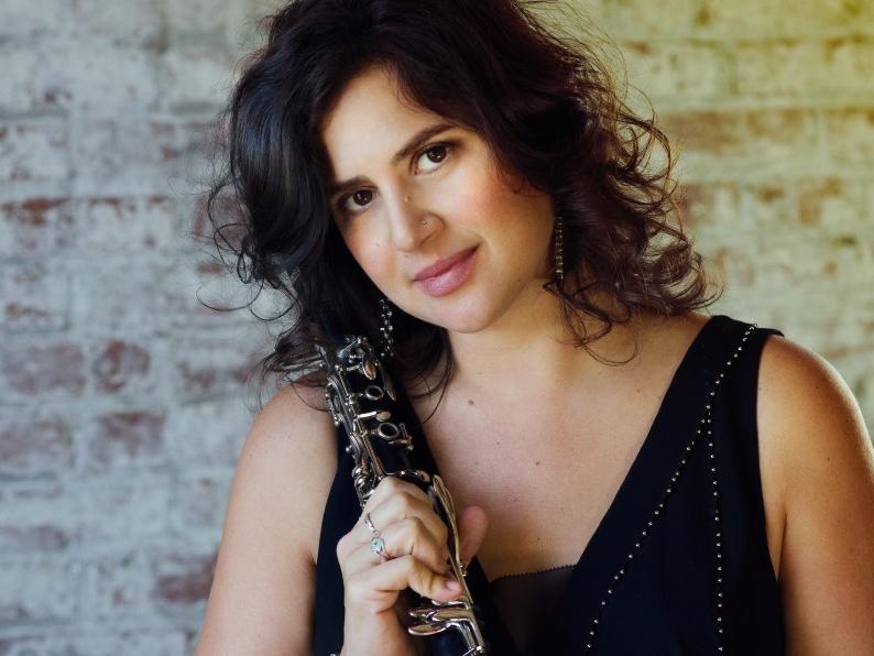 Anat Cohen, shown from the shoulders up, holds a clarinet while she smiles for the camera.