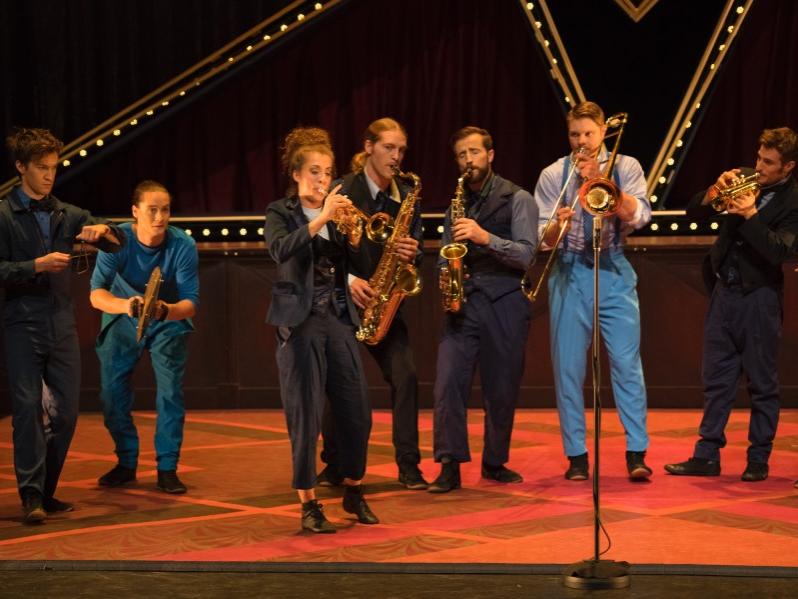 A group of musicians stand in a line on stage facing the audience and play a variety of instruments.