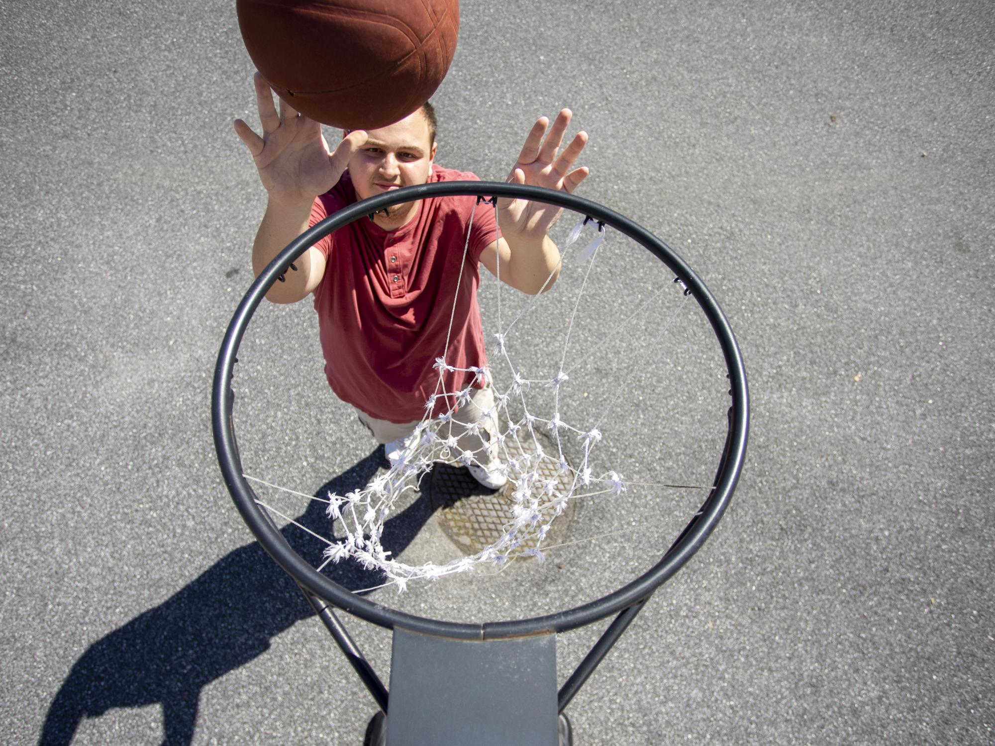 Man in red shirt, arms outstretched above head shooting an orange basketball toward a metal hoop while playing in his driveway.