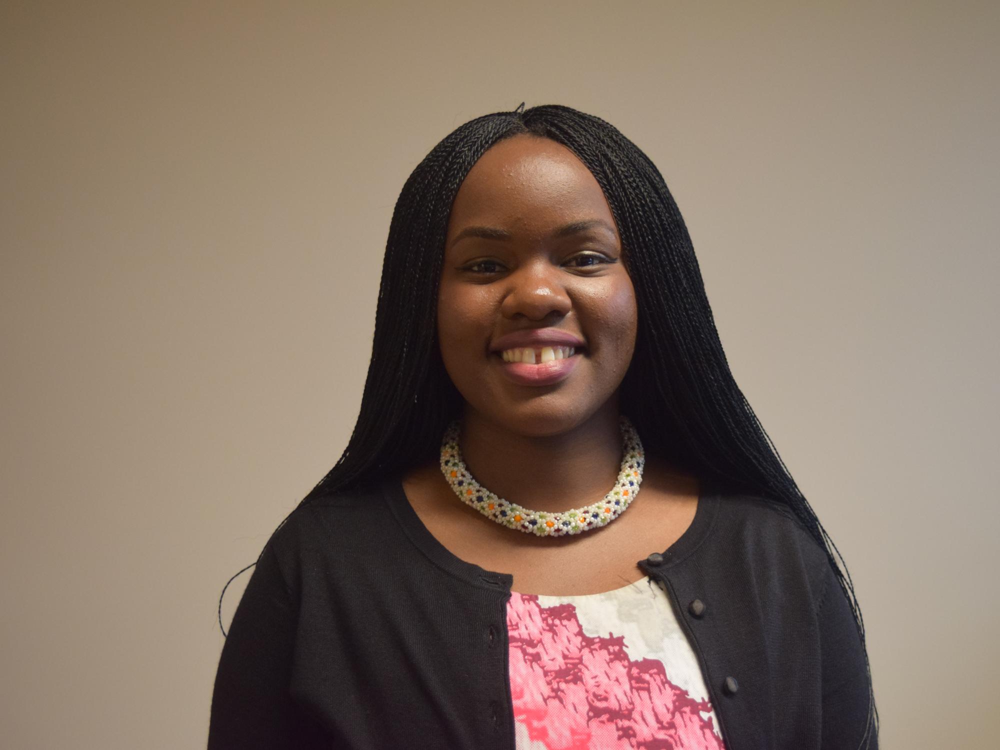 A young woman with long black. braided hair smiles in front of a brown wall. She is wearing a multi-colored beaded necklace, a black sweater and a pink and white  blouse.