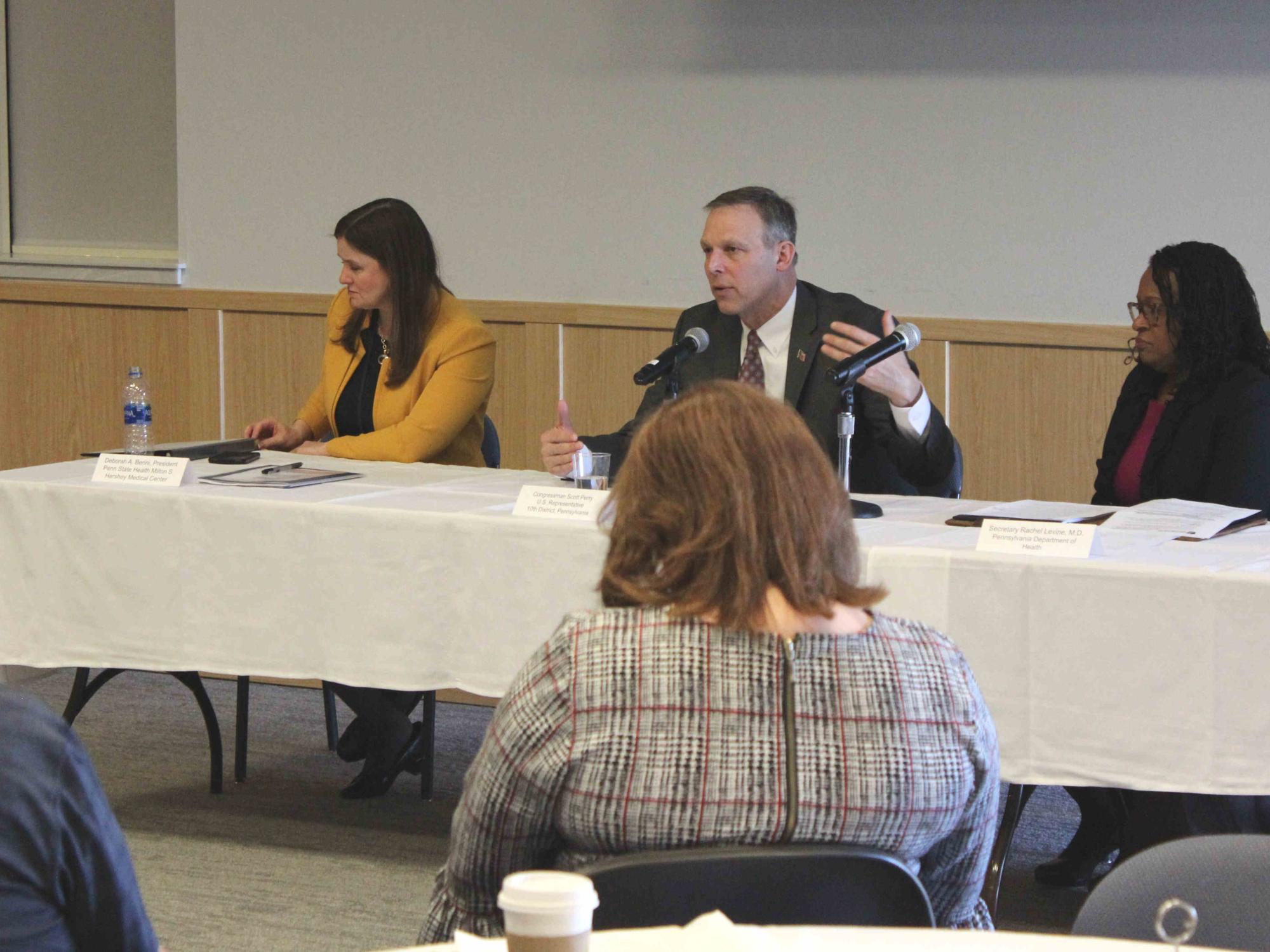 Three people – two women and a man – are seated at a table, facing an audience. The man seated in the middle, is speaking and gesturing with his hand.
