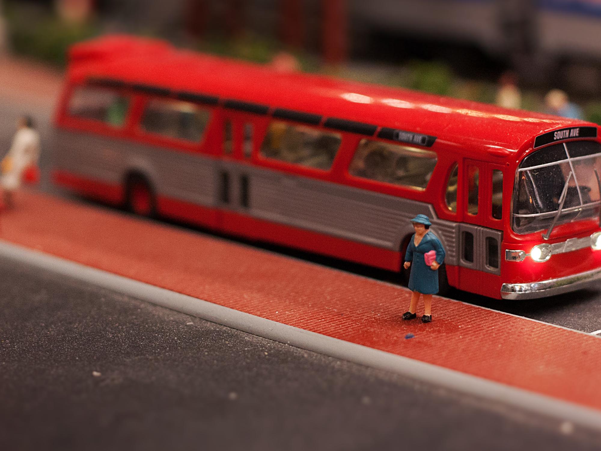 A miniature patron waits by a red bus 