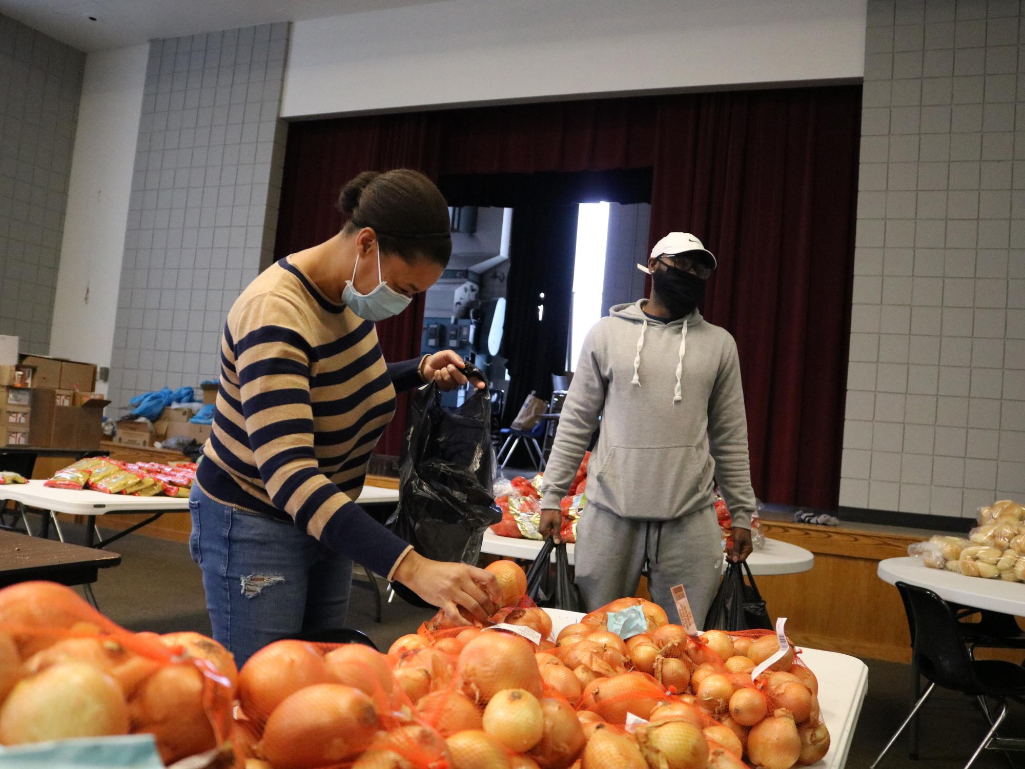 Woman and Man holding plastic bags. Bundles of White Onions sit in front of the frame.