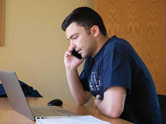 Zach Sowa holds a phone up to his ear while looking at his laptop screen.