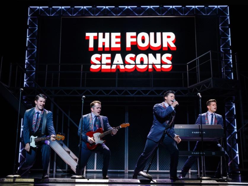 Four men dressed in identical suits and all facing to their left perform their instruments while one performer sings.