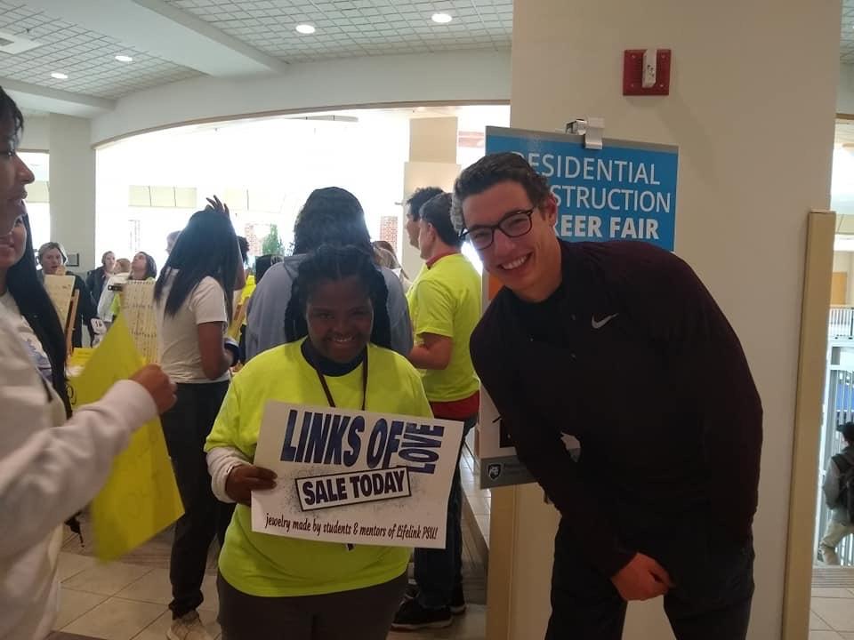 A young man wearing glasses stands with a young woman holding a "Links of Love" sale sign for Lifelink PSU.