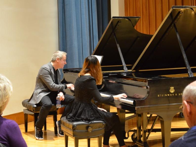 A man and woman each sit at their own piano while he coaches the student musician.
