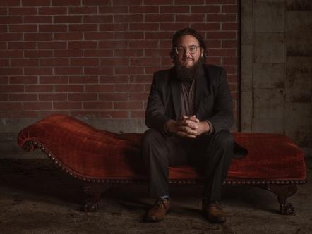 A man sits on a couch with his hands folded and resting on his knees.