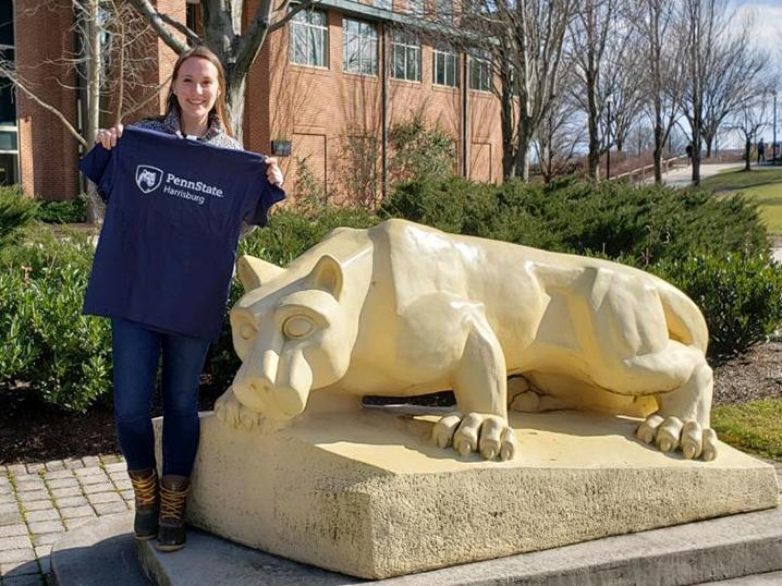 Kailee Shotto stands next to the Harrisburg Lion Shrine and holds up a Penn State Harrisburg tee shirt.