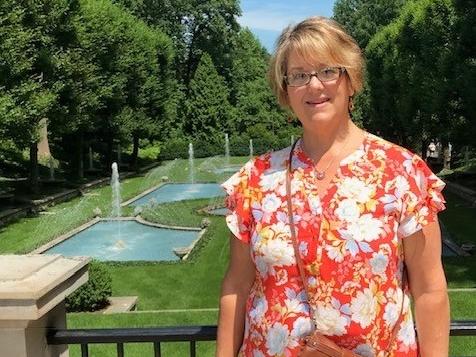 A woman wearing and orange blouse with a white flower pattern stands in front of railing overlooking green grass and fountains