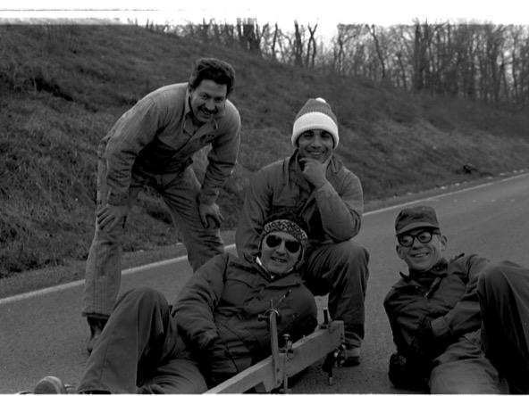 four men standing, kneeling and lying on a road surface