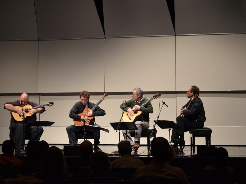 Four guitarists sit in chairs and perform on stage.