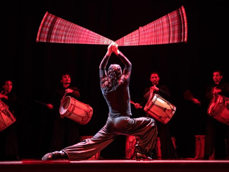 A dancer flicks a leather rope with weighted stones in a circle above his head while other dancers watch.