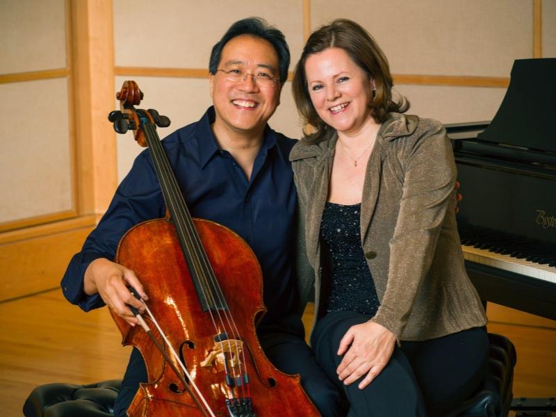 Yo-Yo Ma smiles while cradling his cello in his right arm and his left arm postured around a smiling Kathryn Stott.