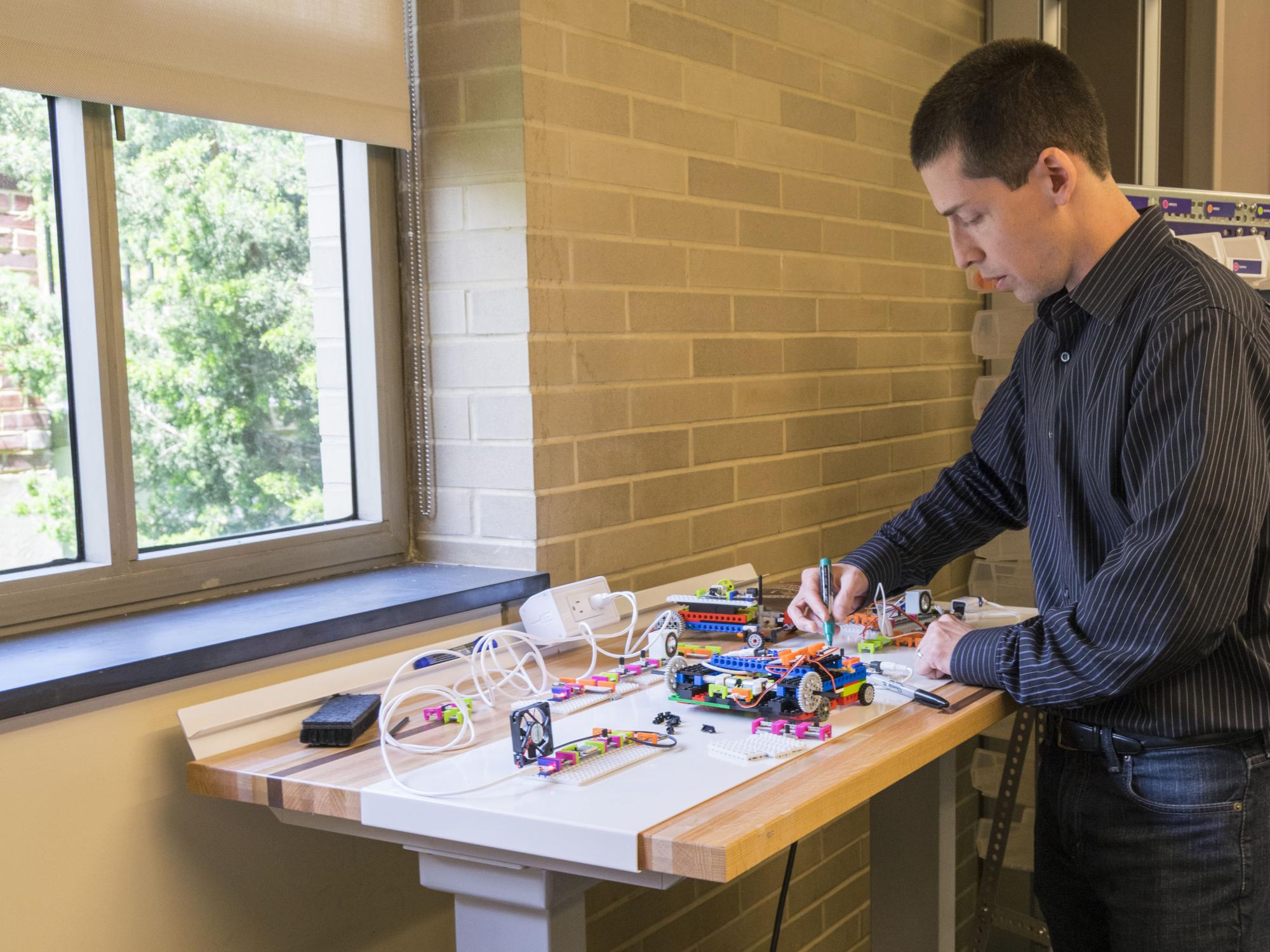 A man in a black outfit stands at a table of littleBits and uses a dry erase marker to write on the table.