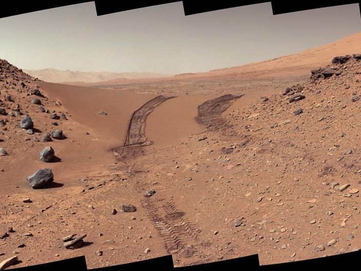 view of Martian landscape, redish brown dirt with occasional rocks showing the tire tracks from the rover