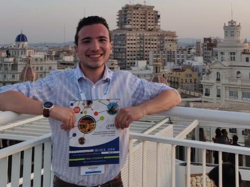 Man standing on an overlook holding an event program with a city behind him. 