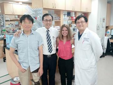 Megan Litz is pictured with three scientists. She wears a pink shirt, a man on the left has a striped shirt, the man to the right of him has a white shirt and a striped shirt, and the other man has a white lab coat on. They stand in a lab.