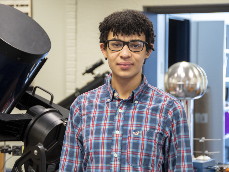 Student wearing glasses and a plaid shirt standing in front of a telescope and other physics-related lab equipment.
