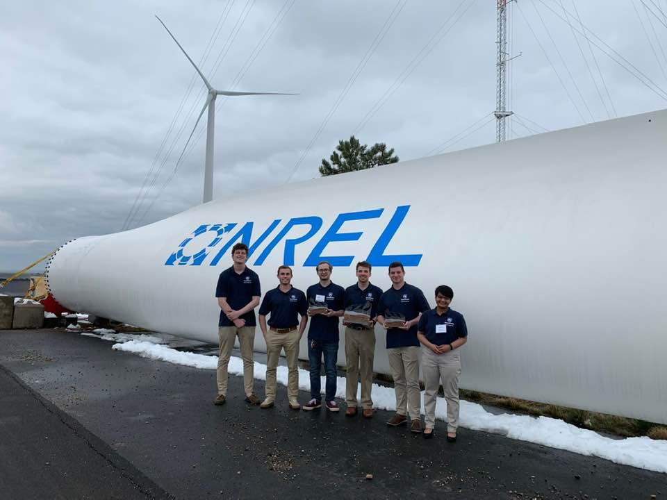 Six students pose with trophies next to a wind turbine blade