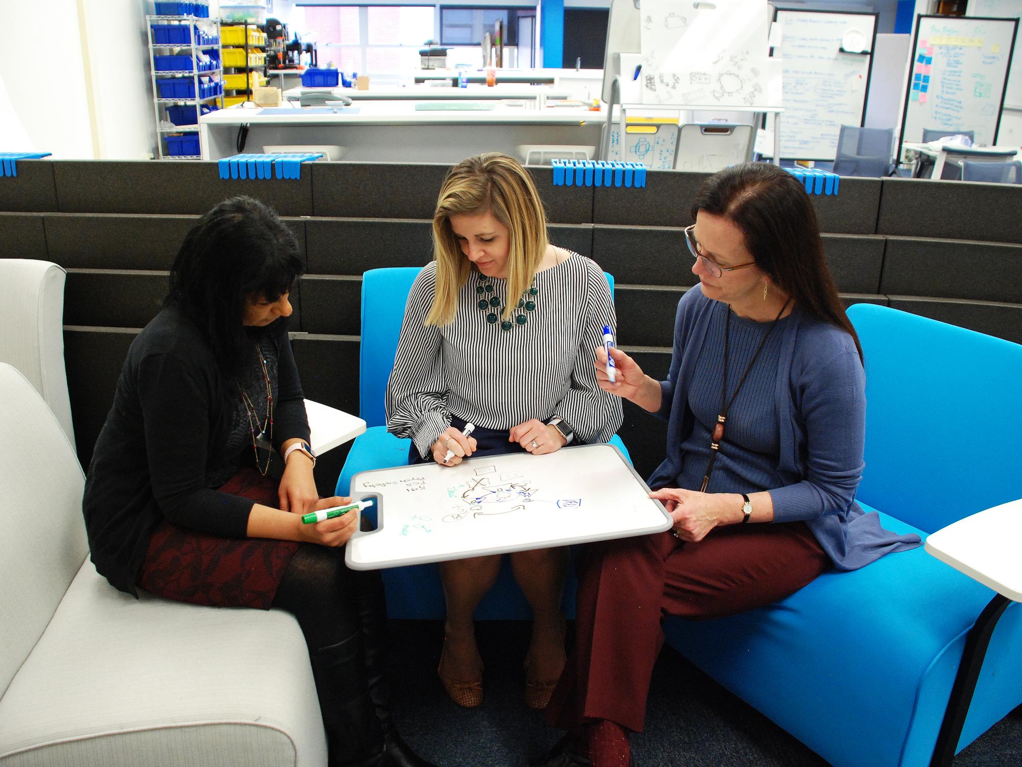 Three women sit in chairs and write on a lap whiteboard