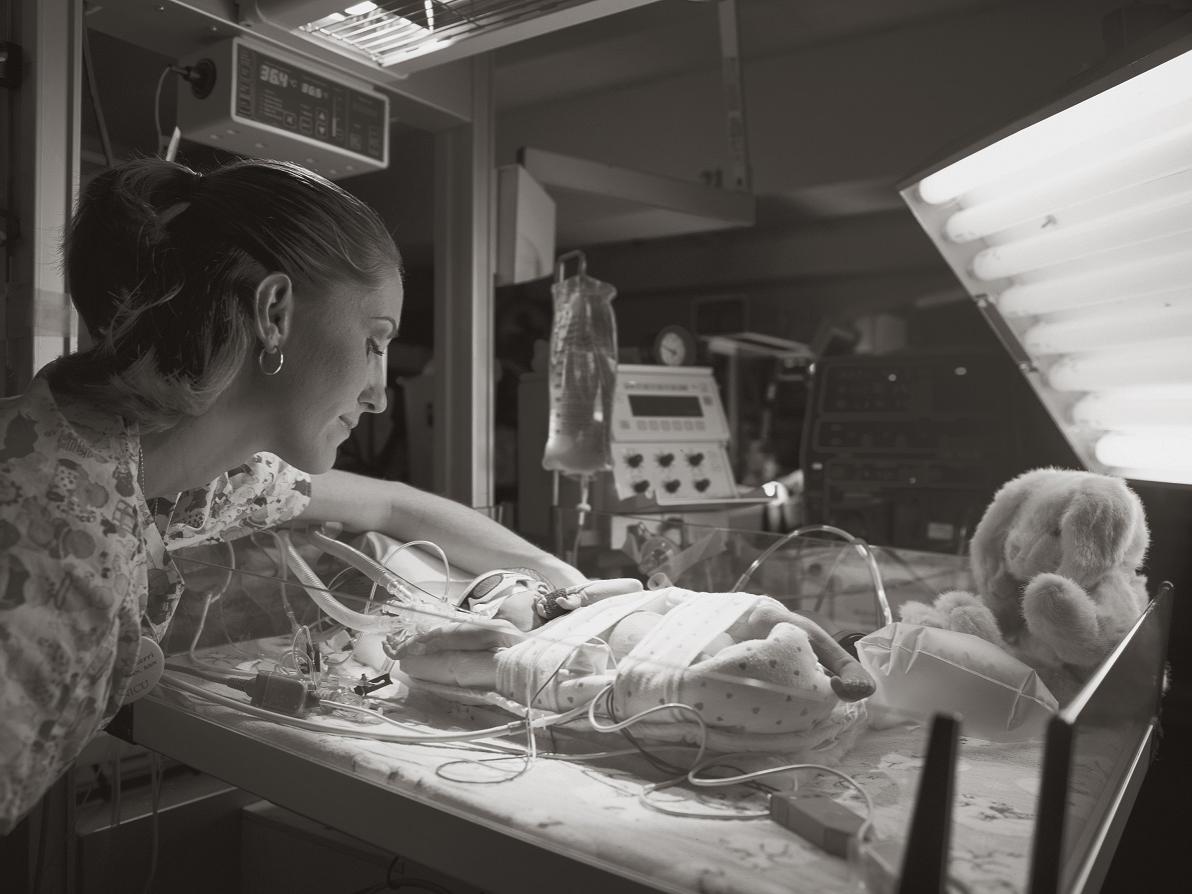 A nurse, wearing scrubs patterned with cartoon pictures of infants, looks over the bassinet of a baby in the Neonatal Intensive Care Unit. Tubing runs from equipment in the background to the baby, who is lying on his back.