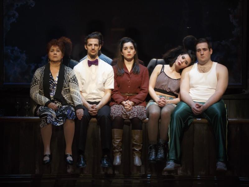 Cast members from the musical “Once” sit on a rail with their legs dangling. 