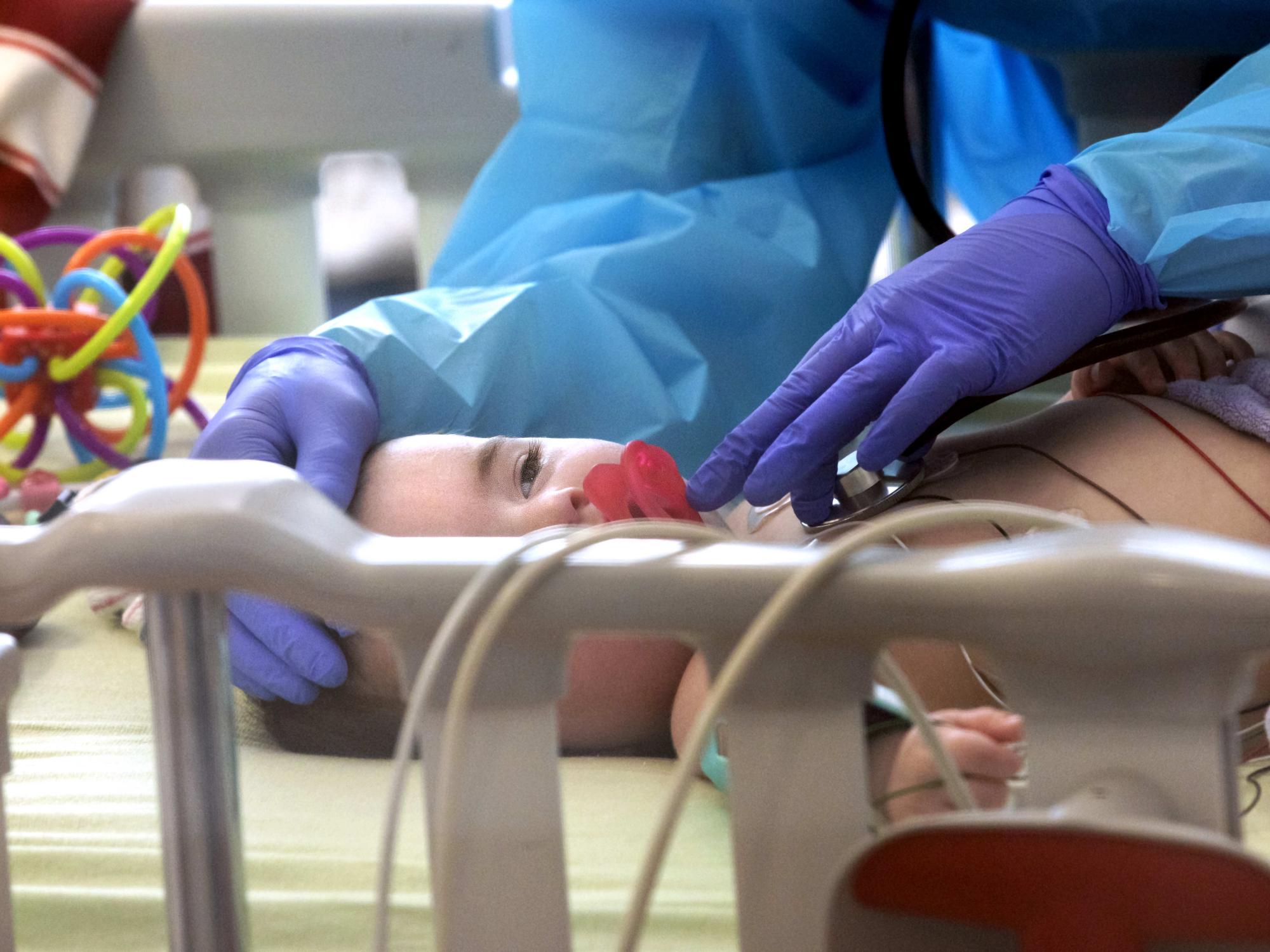 The rubber gloves and gown of a health care worker can be seen holding the end of a stethoscope to the chest of a baby. The baby has a pacifier and stares ahead with half-lidded eyes.