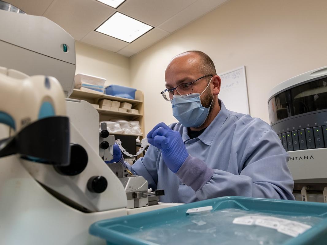 Brandon Borrell is shown wearing personal protective equipment working on a small, paperlike object in front of equipment.