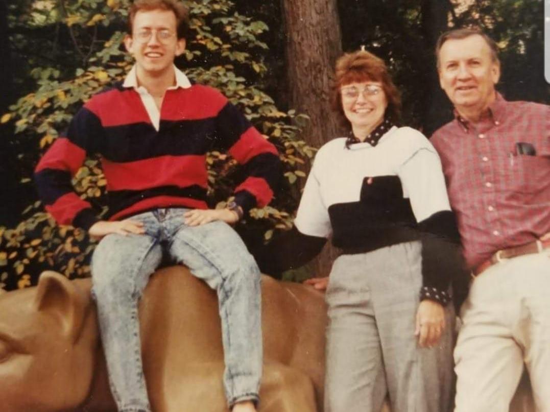 A man sits on the Penn State lion statue with his parents standing beside him.