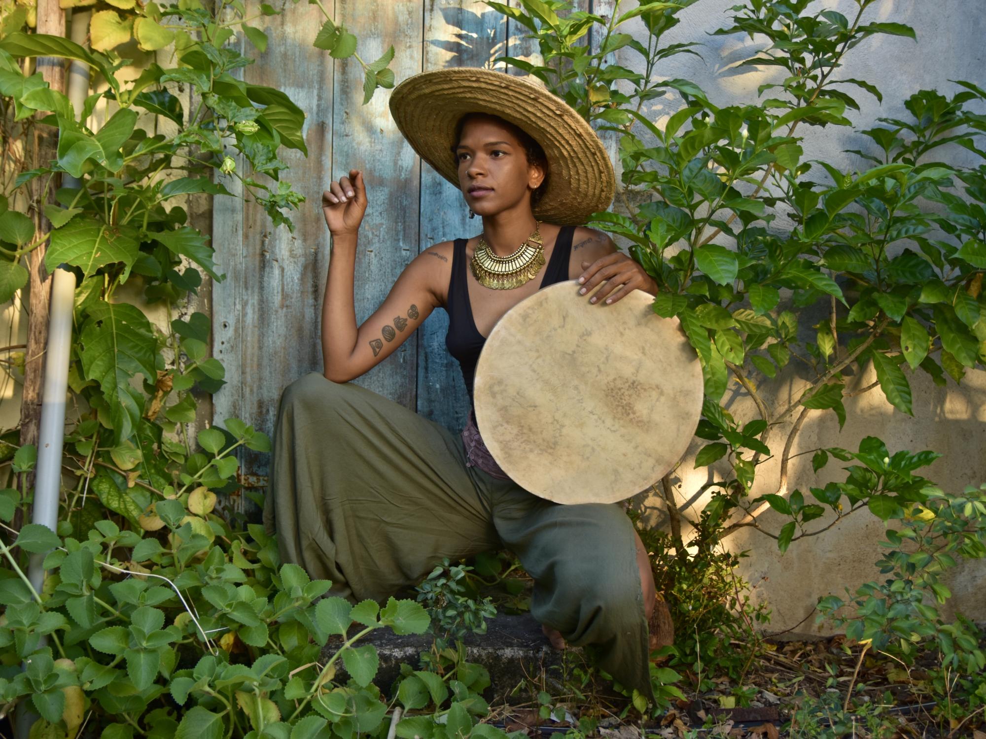 A woman stands amid a lush forest background and holds a drum.