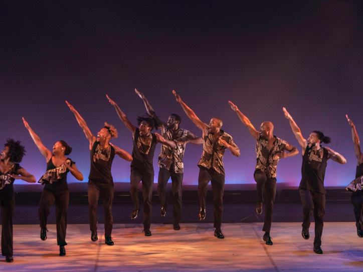 Dancers standing in a line on stage stomp their right feet and extend their right arms into the air.
