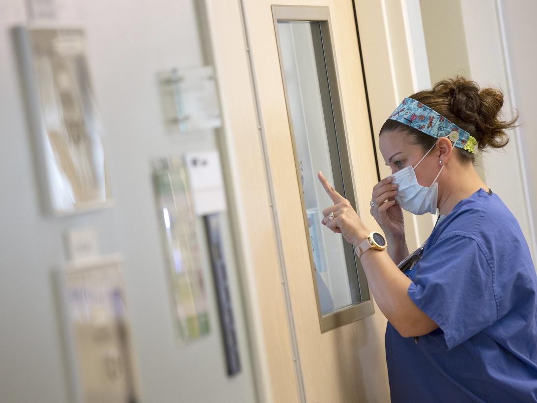 Karen Palladino, wearing scrubs and a surgical mask, presses an index finger against a pane of glass.
